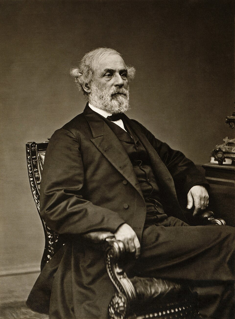 A seated studio portrait of an elderly man with a full white beard in a dark civilian coat, one hand resting on a table.