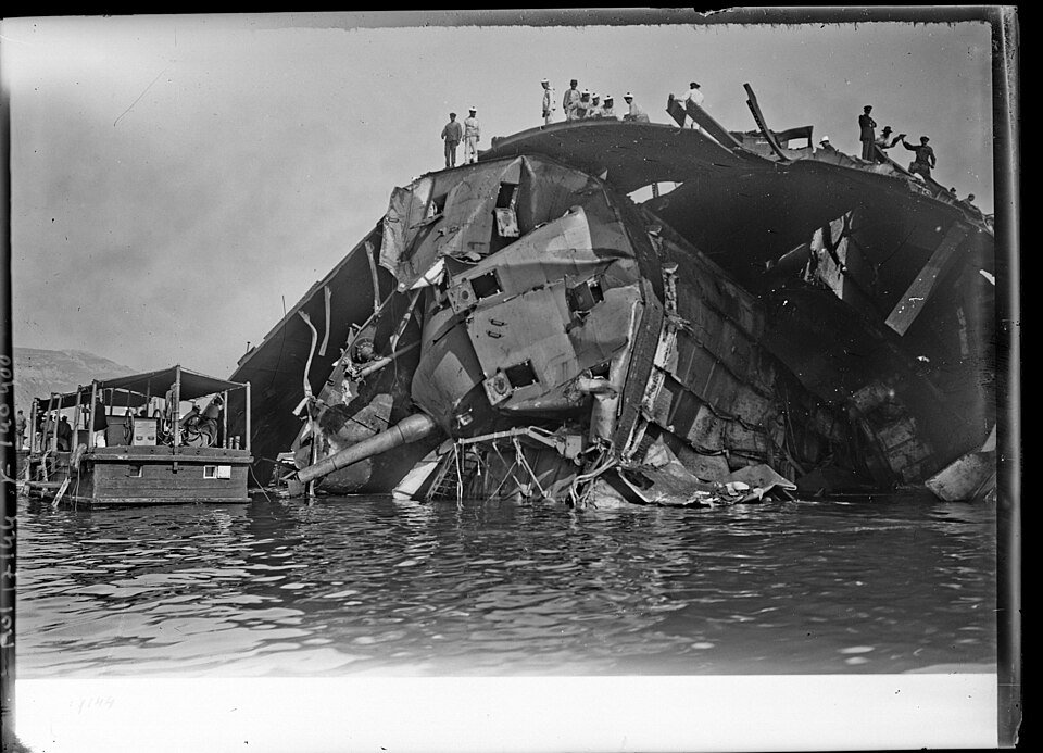 The gutted hull of a large warship lying in harbour, its plating peeled open, with sailors standing on the wreck.