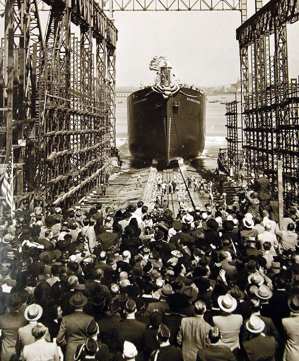 A cargo ship sliding stern-first down a slipway between shipyard gantries as a large crowd in hats watches from below.