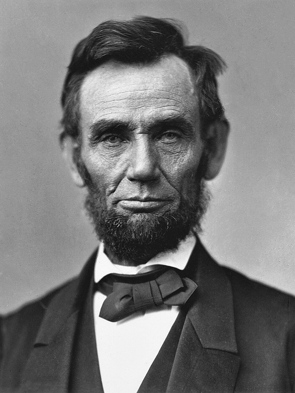A photographic portrait of a gaunt bearded man in a dark suit and bow tie, hair swept back, looking slightly away from the camera.