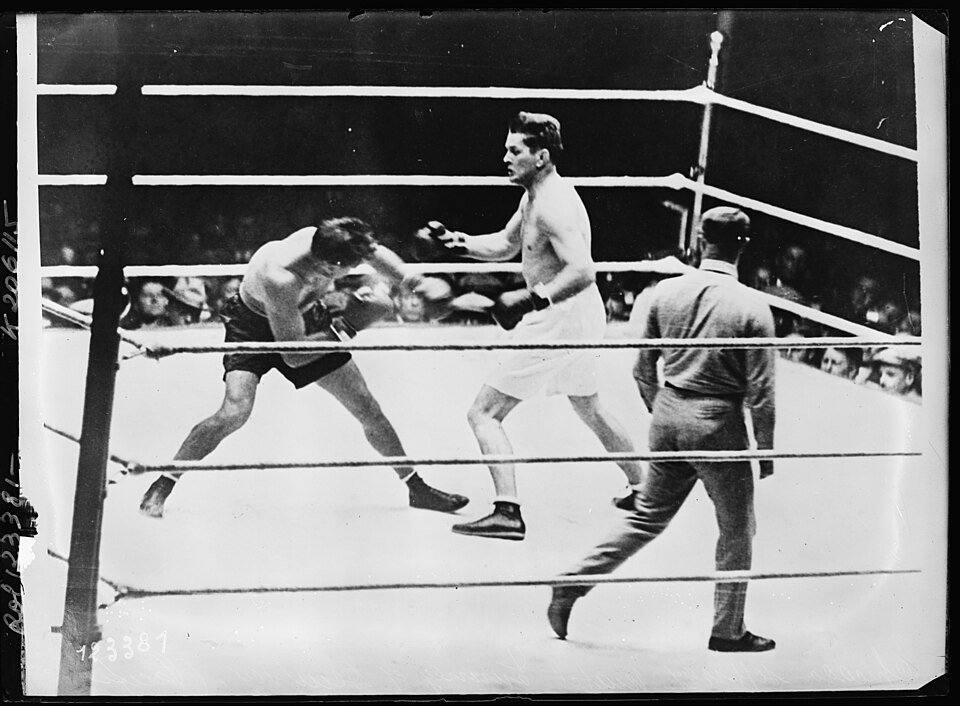 Two boxers in a ring in a crowded arena, one advancing with his guard up while a referee in a suit watches from the corner.