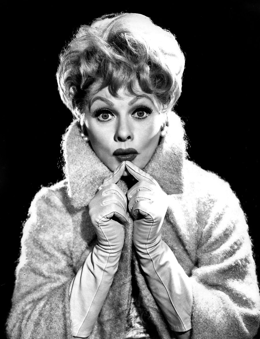 A black and white publicity portrait of a woman with light curled hair in a fur wrap and long gloves, hands clasped near her chin, eyes wide.