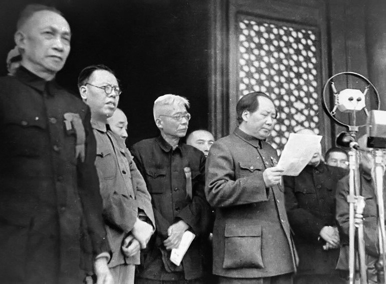 A group of men in plain military tunics standing on a balcony, one reading from a sheet of paper into a large microphone.