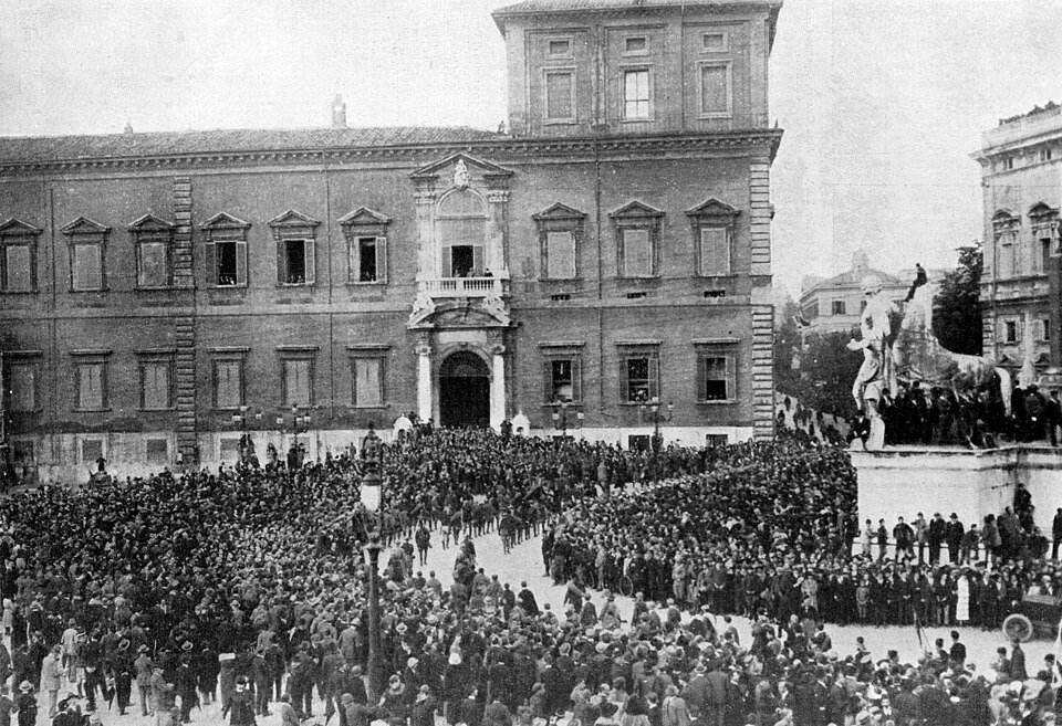A vast crowd packed into a square in front of a long classical palace, with a monument and figures on a balcony above them.