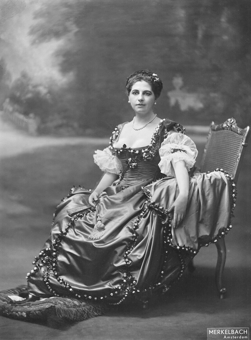 A studio photograph of a woman in an elaborate embroidered gown and headdress reclining on a chaise longue, looking directly at the camera.