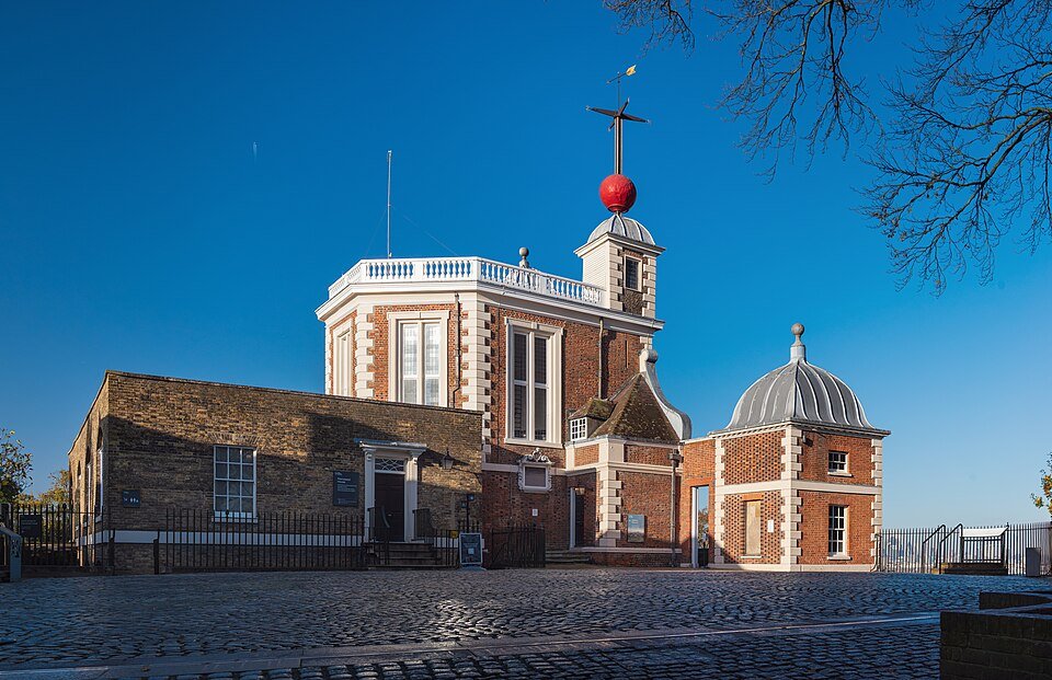 A brick and stone observatory building with white quoins and a small turret carrying a tall mast topped by a large red ball, photographed against a deep blue sky.