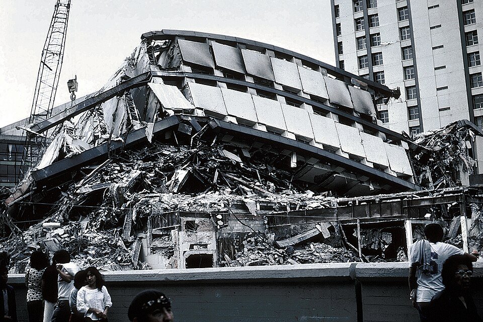 A modern office block collapsed into a folded heap of concrete and metal panels, with a crane behind and onlookers in front.