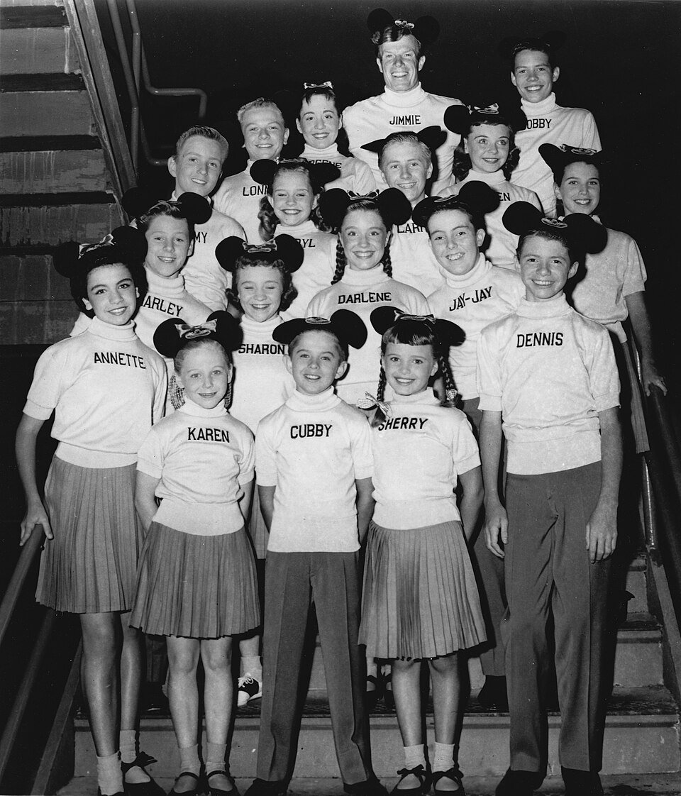 A group of children and teenagers posed on a staircase wearing matching white shirts with their first names printed on them and black mouse-ear caps.