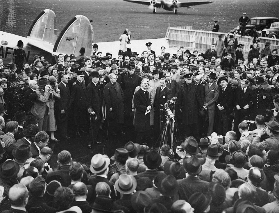 A man in a dark overcoat holding up a sheet of paper before a bank of microphones, surrounded by a large crowd on an airfield with an airliner behind.