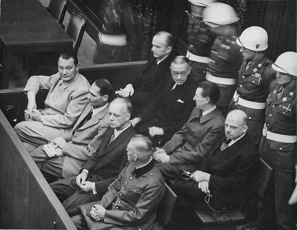 Rows of men in suits and uniforms sitting on wooden benches in a courtroom dock, several wearing headphones, with helmeted military police standing behind them.