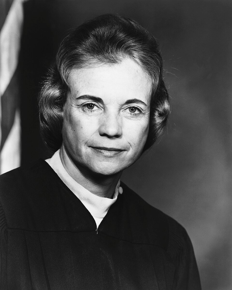 A formal portrait of a woman in black judicial robes in front of a flag.