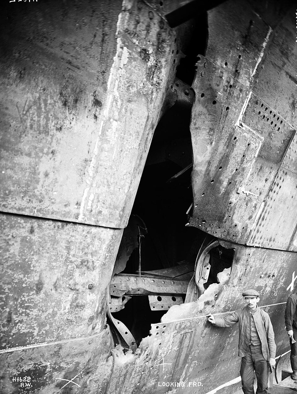 A photograph of a huge riveted steel hull torn open, with a workman in a flat cap standing beside the hole for scale.