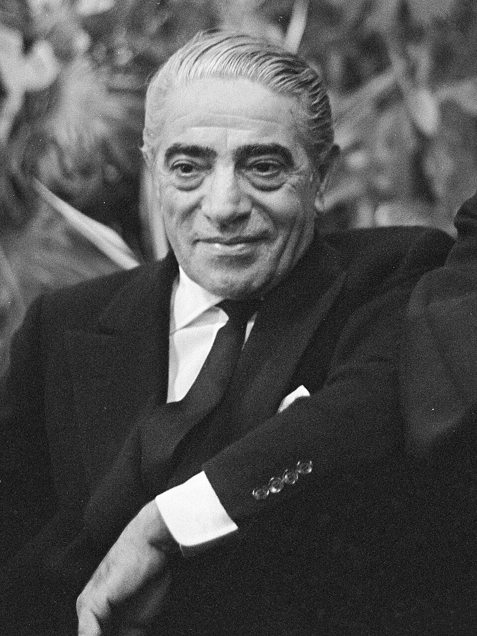 A black and white portrait of a tanned middle-aged man with slicked-back grey hair in a dark suit and tie, smiling slightly at the camera.