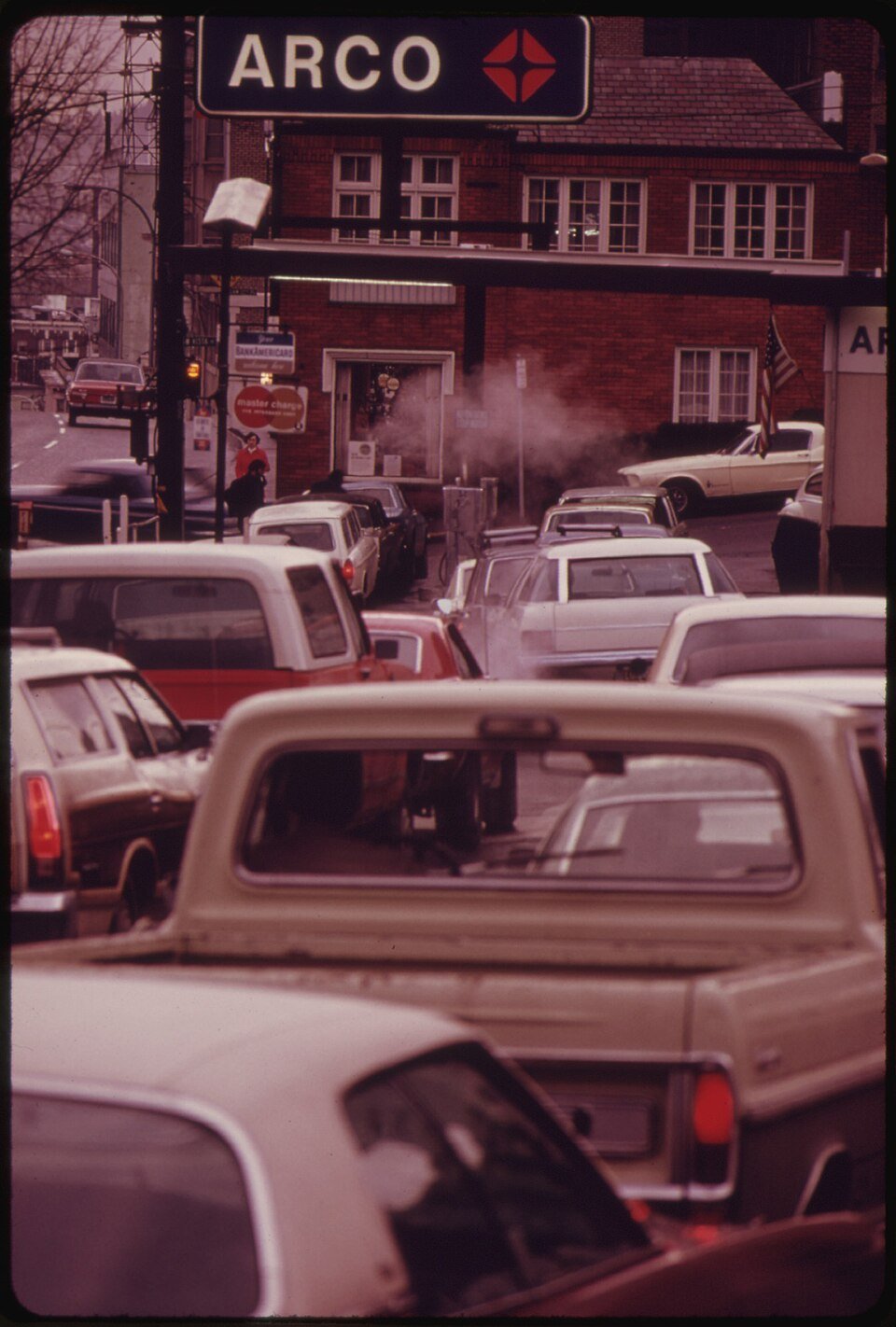 A colour photograph looking down a long queue of 1970s cars and pickup trucks waiting bumper to bumper at a petrol station, a tall illuminated sign overhead.