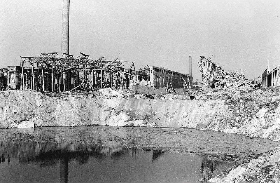 A black and white photograph of a vast water-filled crater in front of the twisted steel skeleton of a factory.