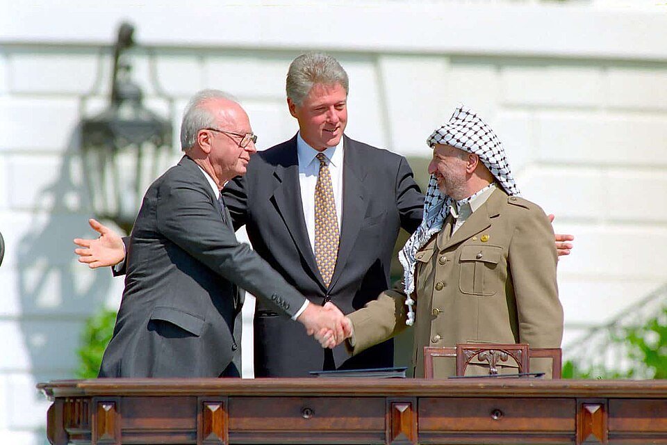 Colour photograph of two older men shaking hands on a lawn while a third stands between them with arms outstretched.