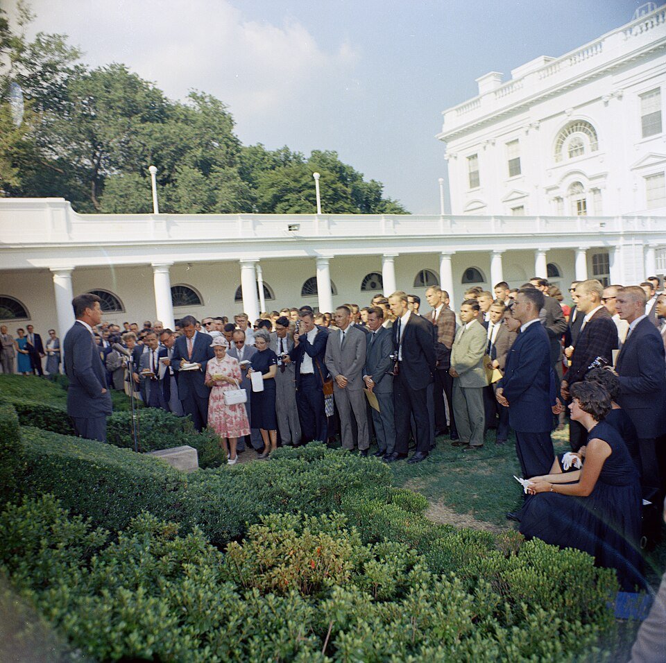A colour photograph of a man in a dark suit addressing a large crowd of young people in a formal garden beside a white colonnaded building.