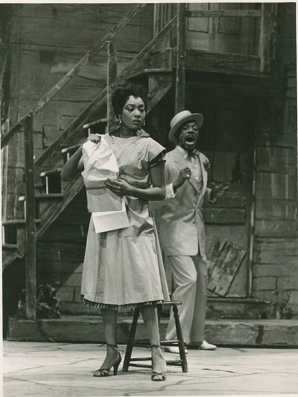 A stage photograph of a woman in a plain dress holding a crumpled paper, with a man in a light suit and straw hat singing behind her, in front of a tenement set with wooden stairs.