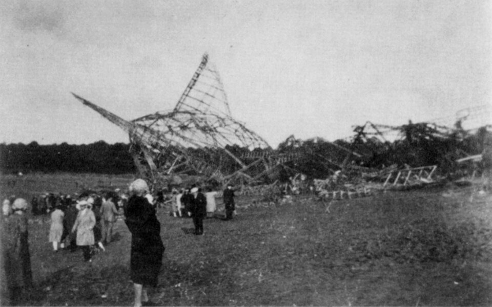 The stripped metal skeleton of a huge airship lying broken across a field, with a small crowd of onlookers in coats and hats standing in front of it.