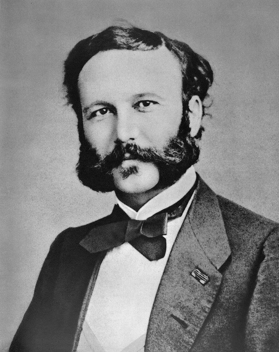 A photographic portrait of a young man with a full dark beard and moustache in a heavy jacket and bow tie, looking slightly past the camera.