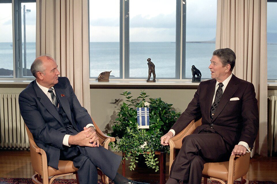 Two men in dark suits sitting in armchairs facing each other in a bright room, a wide window behind them showing grey sea and cloud.