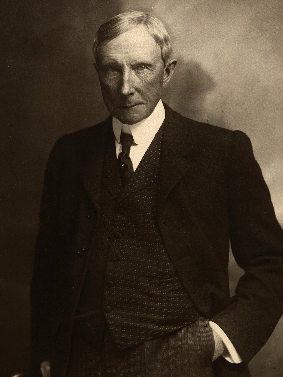 A photograph of a gaunt elderly man in a high collar and dark coat, looking directly at the camera.