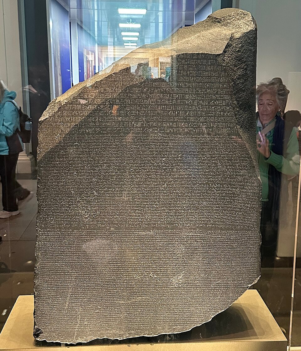 A large slab of dark grey stone covered in three bands of different scripts, displayed in a glass case.