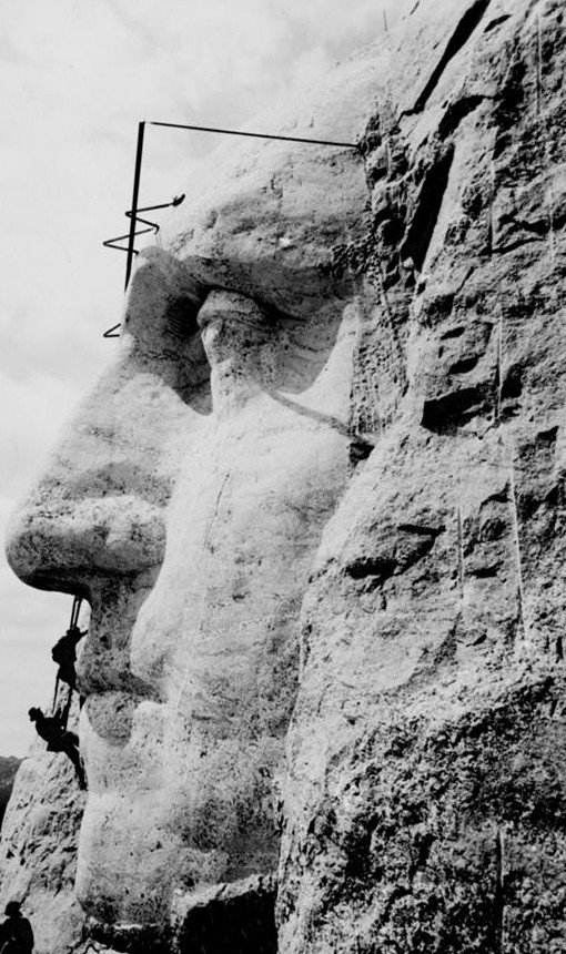 A rough-hewn colossal face emerging from a granite cliff, with steel scaffolding above it and two tiny workmen suspended on ropes beside the nose.