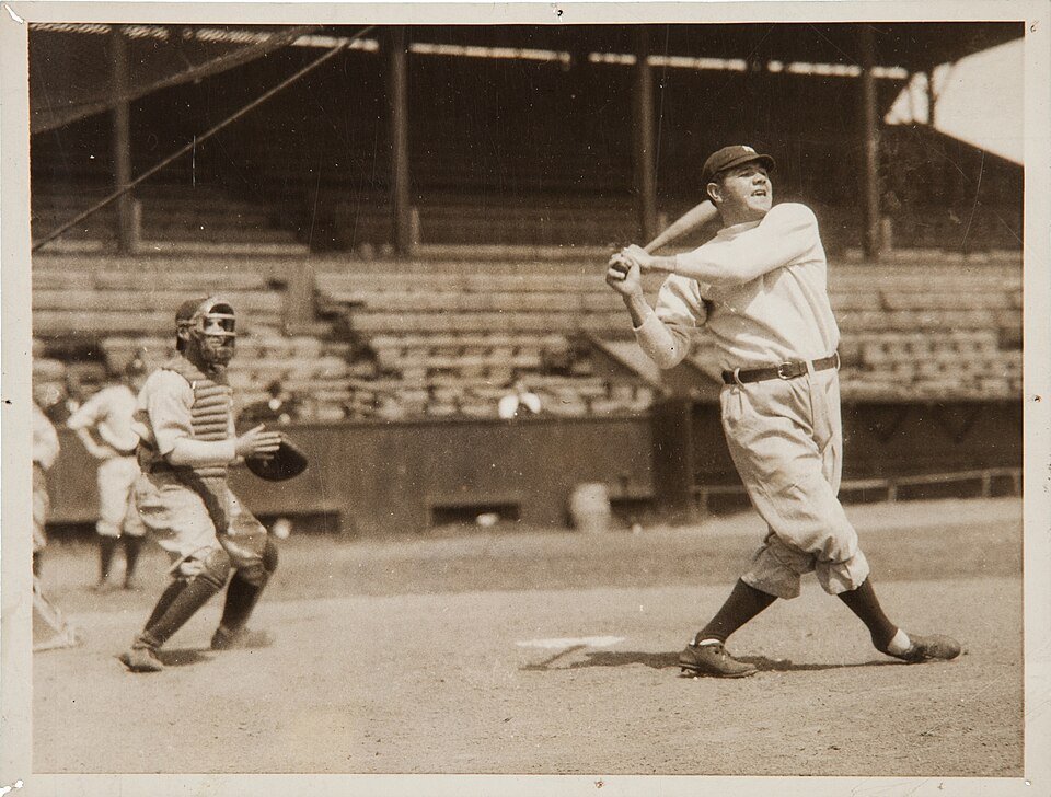 A baseball player in a pale uniform following through on a full swing, the catcher and empty stands behind him.