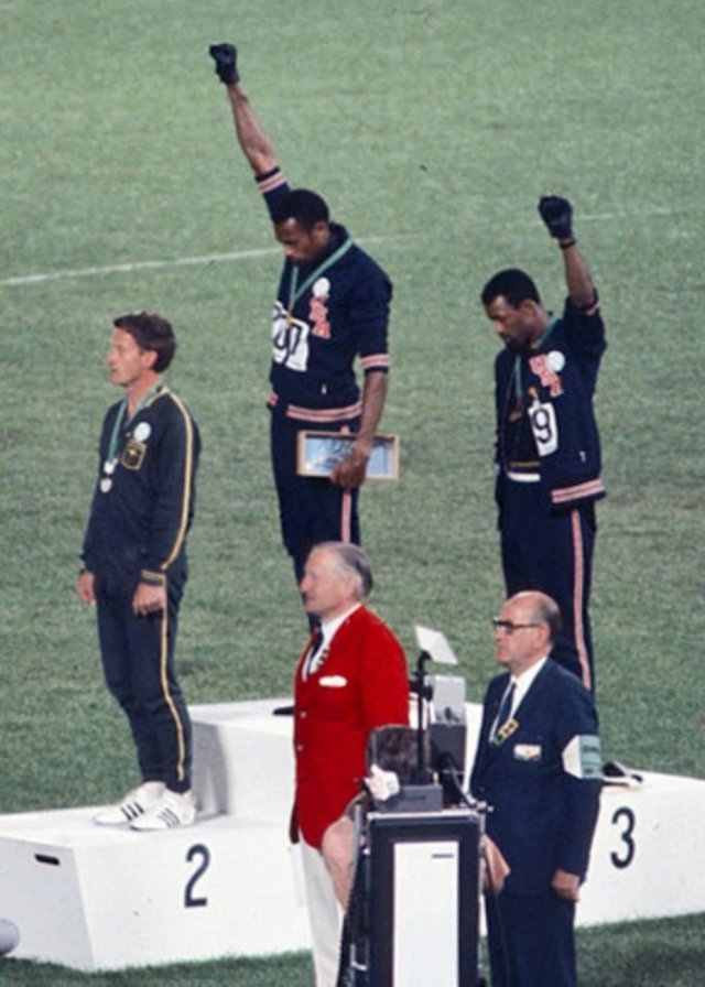 A colour photograph of two athletes on an Olympic podium with heads bowed and black-gloved fists raised, a third athlete standing still on the step below them.