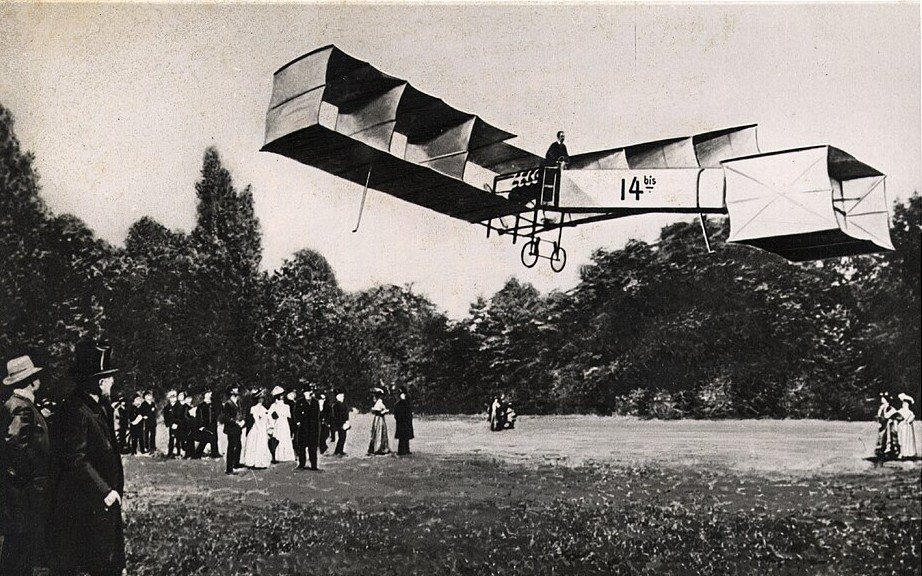 An early aeroplane built of box-kite panels flies low over open grass while a crowd of men in hats and women in long dresses watches from the edge of the field.