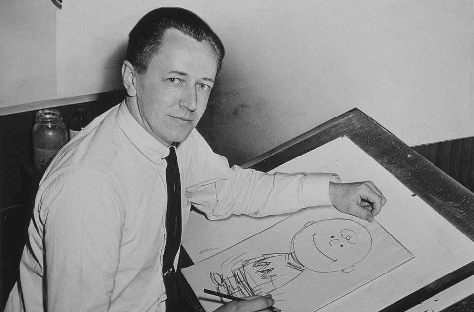 A man in shirtsleeves and a tie sitting at a drawing board, looking at the camera over his shoulder, a pencil sketch of a round-headed boy on the paper in front of him.