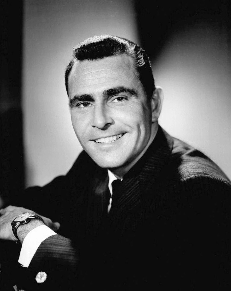 A studio portrait of a man in a dark jacket smiling at the camera, one arm resting in front of him, a wristwatch visible.