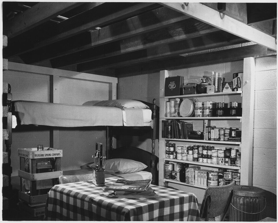 A basement room with bunk beds, a chequered tablecloth, shelves stacked with tinned food, a first aid box and a metal bin, lit from above by exposed floor joists.