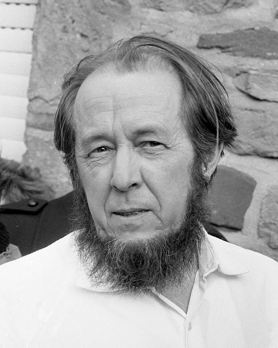 A black and white portrait of a middle-aged man with a receding hairline and a full beard, wearing an open-necked white shirt, standing in front of a stone wall.
