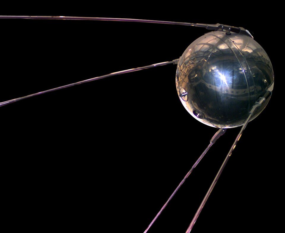 A polished metal sphere with four long thin antennae trailing back from it, photographed against a black background.
