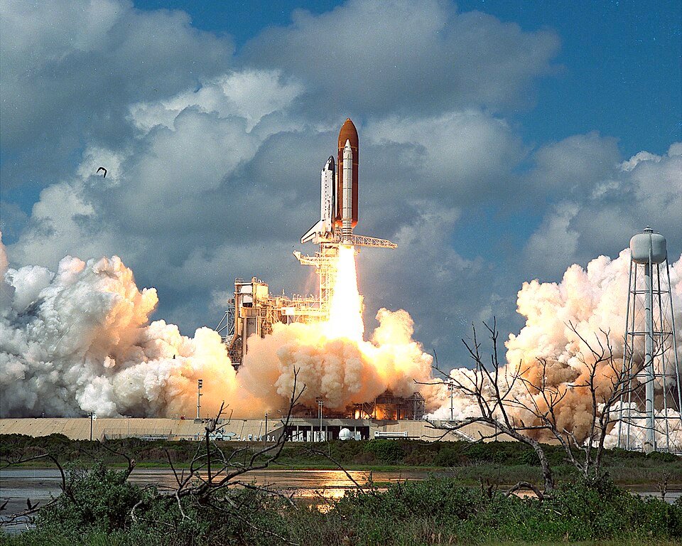 A space shuttle climbing on a column of flame and smoke against a blue sky.