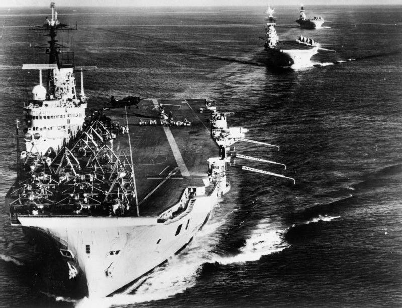 Two aircraft carriers photographed from astern steaming in line across open sea, aircraft ranged on the flight deck of the nearer ship.