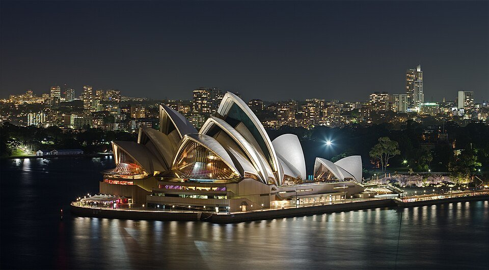 A night photograph of a waterfront building of white curved shells lit from within, reflected in dark harbour water with a city skyline behind.