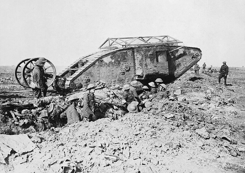 A rhomboid armoured vehicle with a steering tail on wheels, surrounded by soldiers in steel helmets in a cratered landscape.