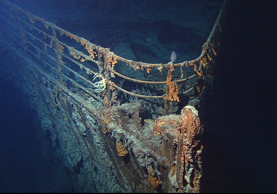 Underwater photograph of a rusted ship bow encrusted with orange growths, railings still intact, lit against deep blue darkness.