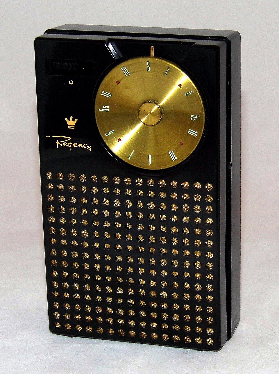 A small black pocket radio standing upright, with a large gold tuning dial at the top and a panel of gold perforations below it.