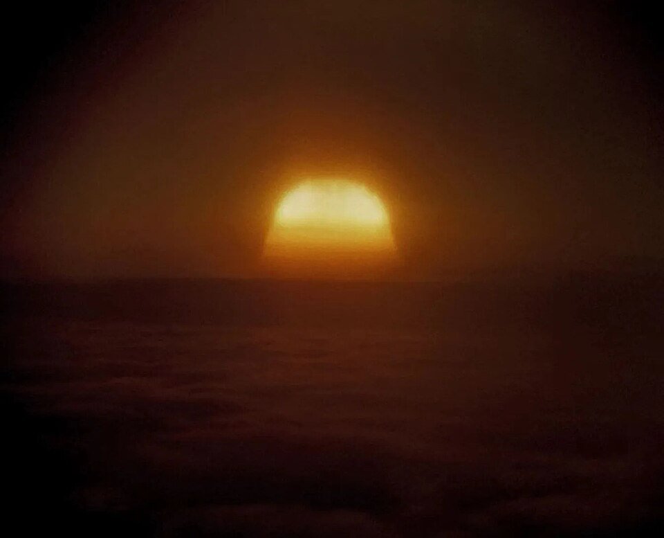 A vast dull orange fireball sitting on a dark horizon under a black sky, its edges blurred into haze.