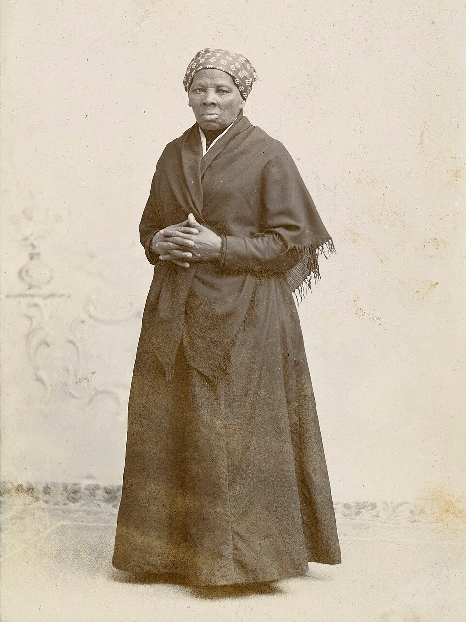 A sepia studio photograph of a woman in a long dark dress and patterned headscarf standing with her hands clasped.