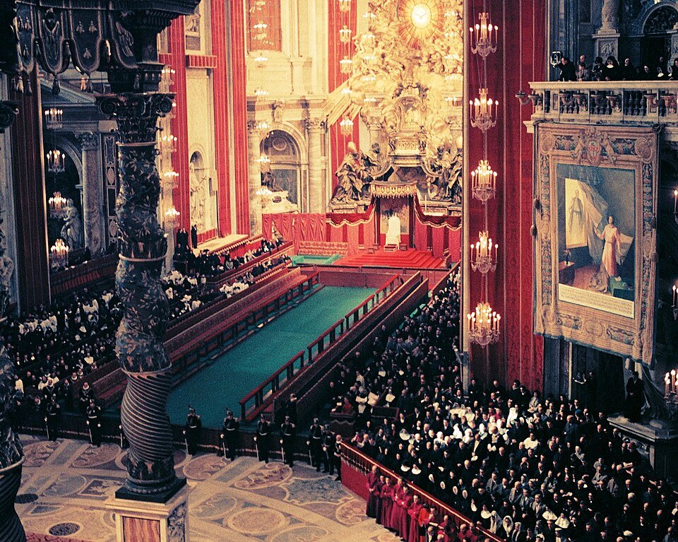 A vast church interior seen from a balcony, with tiers of green-draped benches packed with clergy running down the nave beneath red hangings, chandeliers and a gilded canopy.