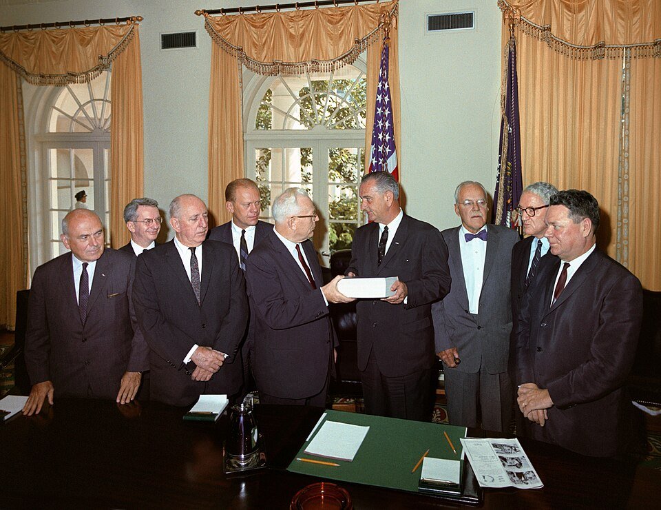 A group of men in dark suits standing in a formal office as one hands a thick bound volume to another.