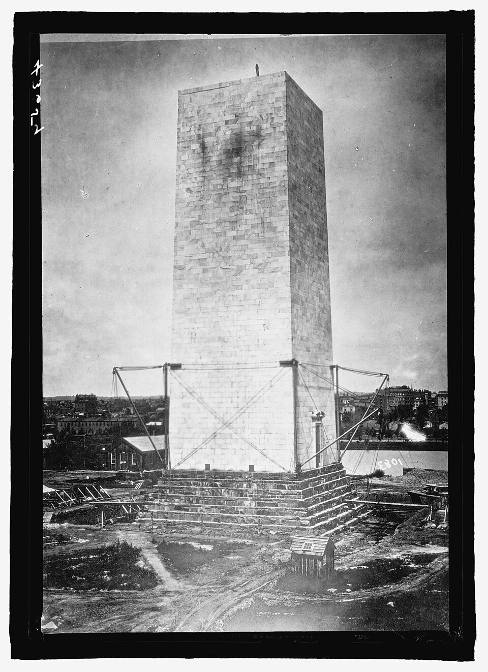 A tall unfinished stone obelisk standing as a squat stump on a bare construction site, braced with iron scaffolding at its base, with a low city skyline behind.