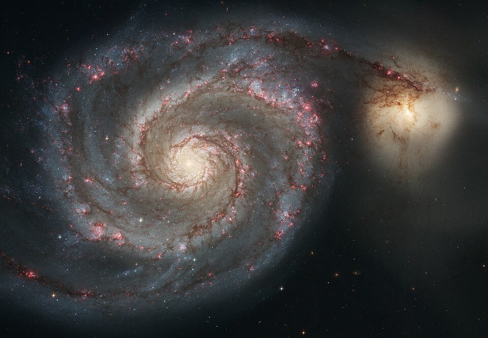A telescope image of a spiral galaxy with pink star-forming knots along its blue arms, a smaller yellow companion galaxy attached at the end of one arm.