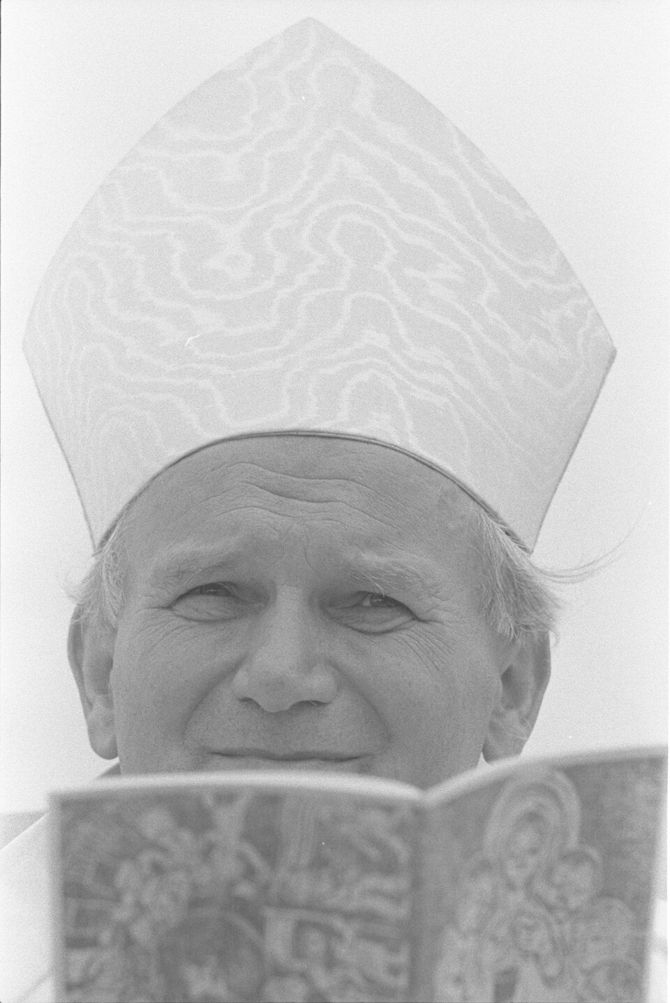 A close-up photograph of a smiling clergyman in a tall embroidered mitre, reading from an illuminated book held up in front of him.