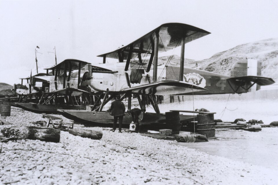 Three biplanes on floats drawn up on a shingle beach beside oil drums, a man in overalls standing among them.